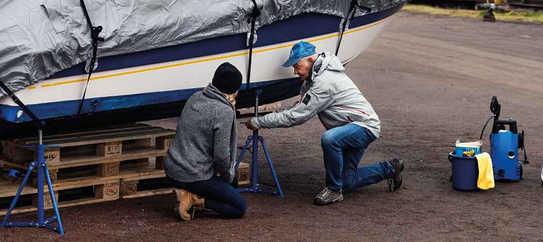 En äldre man i en blå keps, ljusgrå jacka och blåa jeans sitter på knä på marken och spänner upp en  båtstötta. På båtstöttorna står en vit båt med mörkblå botten. Bredvid mannen sitten en kvinna på knä på marken med ryggen mot.