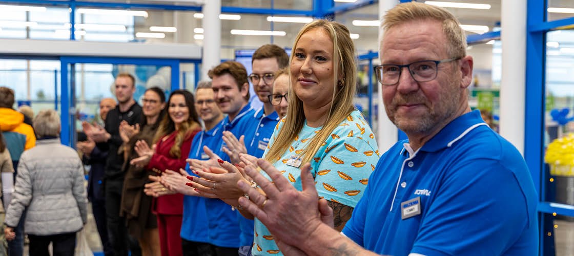 Tre medarbetare i blå Biltema-tröjor står framför en butikshylla med LED-lampor, alla med namnskyltar och ett leende.