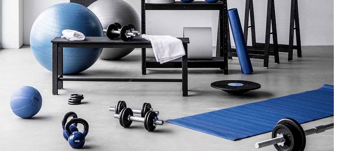 Ljust och välorganiserat hemmagym med träningsutrustning som kettlebells, hantlar, skivstång, träningsbollar och yogamatta. Utrustningen är prydligt placerad på en hylla och på golvet i ett modernt träningsrum med betonggolv och vita väggar. En tavla med ett handskrivet "Squat Challenge"-schema bidrar till träningsmiljön.