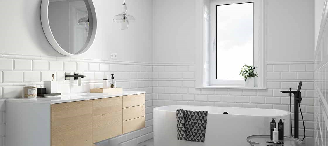 Ett ljust kaklat badrum med ett vitt badkar, en rund väggspegel och en kommod med ljusa trälådor.