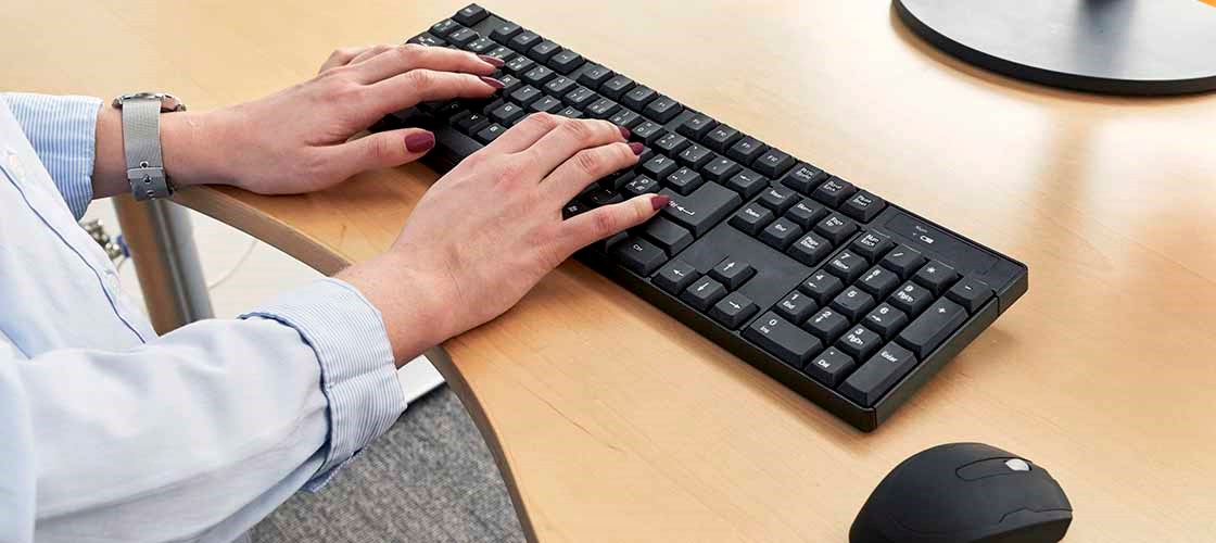 A pair of female hands work on a black keyboard. On her left arm, the woman is wearing a silver watch. On the right, on the table, you can see parts of a computer mouse.