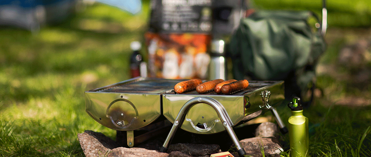 Bärbar campinggrill med korvar som tillagas utomhus på en campingplats.
