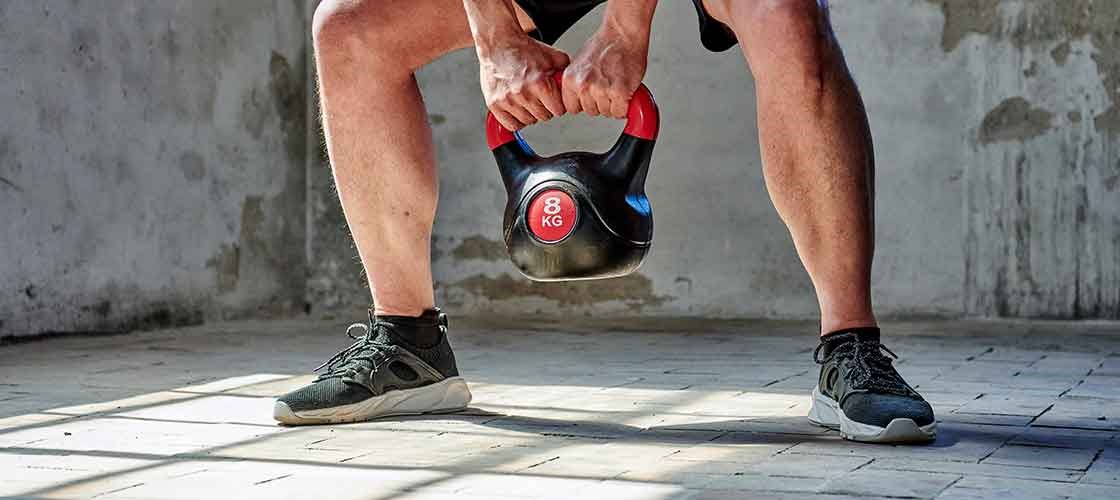 En person i träningskläder lyfter en kettlebell från golvet i ett betongrum med obehandlade väggar.