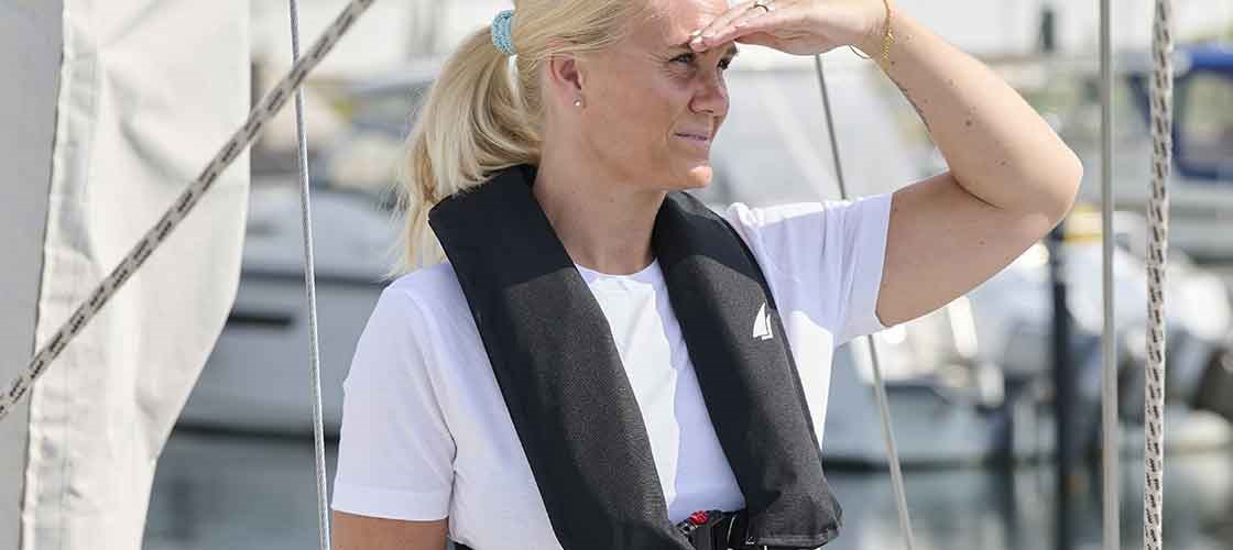 Kvinna som står på däcket på ett segelfartyg iklädd en uppblåsbar flytväst. Används som inspiration för guiden: "Hur du kontrollerar din uppblåsbara flytväst - steg för steg".