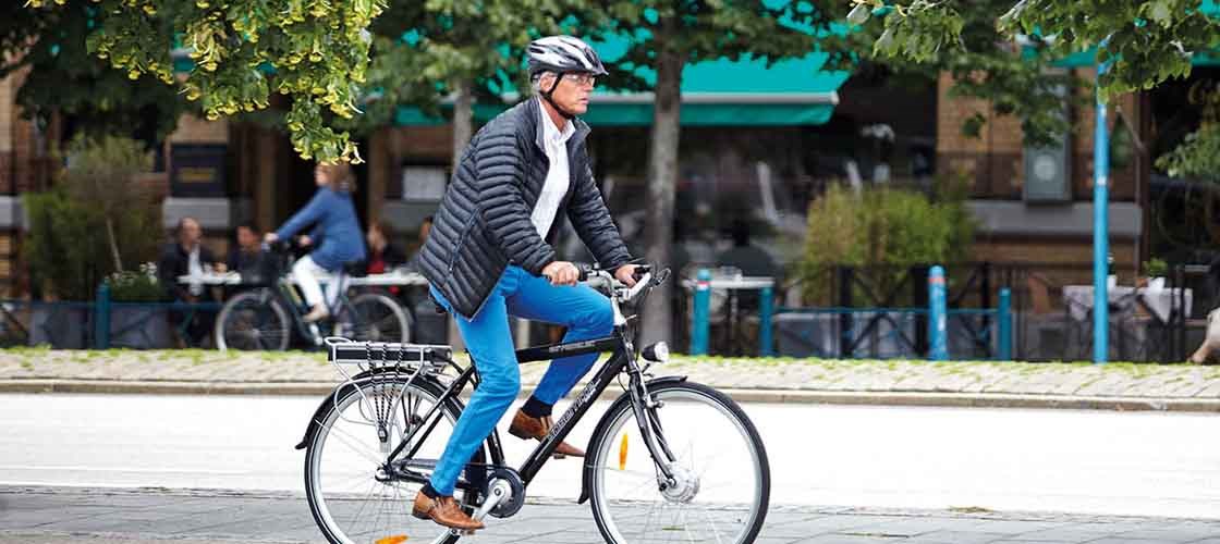 Man med hjälm cyklar på en stadsgata med butiker och träd i bakgrunden.