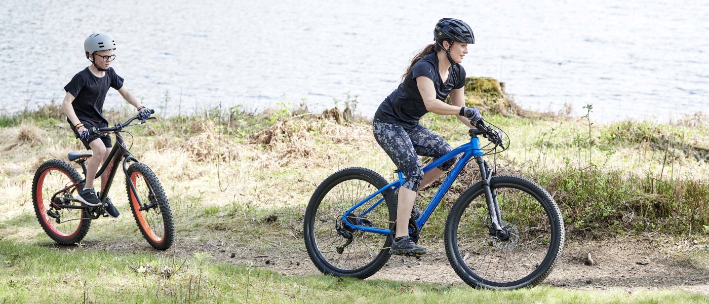 Två personer cyklar på en stig nära vatten och grönska, båda med hjälm och terrängcyklar. Den ena personen färdas på en blå cykel medan den andra följer bakom på en mindre cykel. Sjön och den naturliga omgivningen syns i bakgrunden.