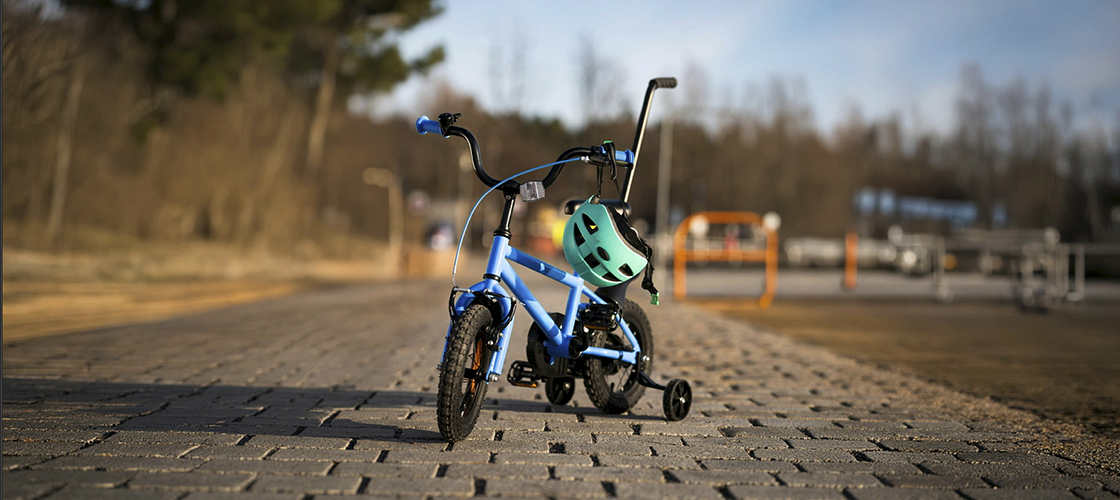 En blå barncykel med stödhjul står parkerad på en stenlagd väg utomhus. En turkos cykelhjälm hänger på styret, vilket förstärker fokus på säker cykling för barn. Bakgrunden är mjukt suddig med träd och omgivande miljö i naturligt dagsljus.