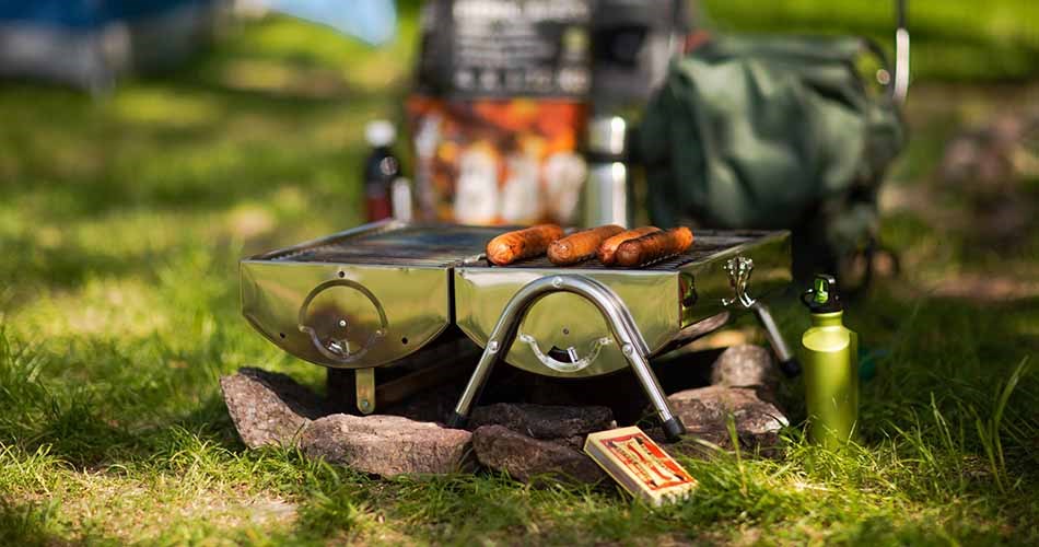 En portabel grill står placerad på marken i ett grönområde och används för att grilla korvar.
Grillen är uppställd på små ben ovanpå stenar och omges av gräs och utrustning för utomhusbruk.
I bakgrunden syns en väska, en flaska och förpackningar som tyder på en utflykt eller camping.
