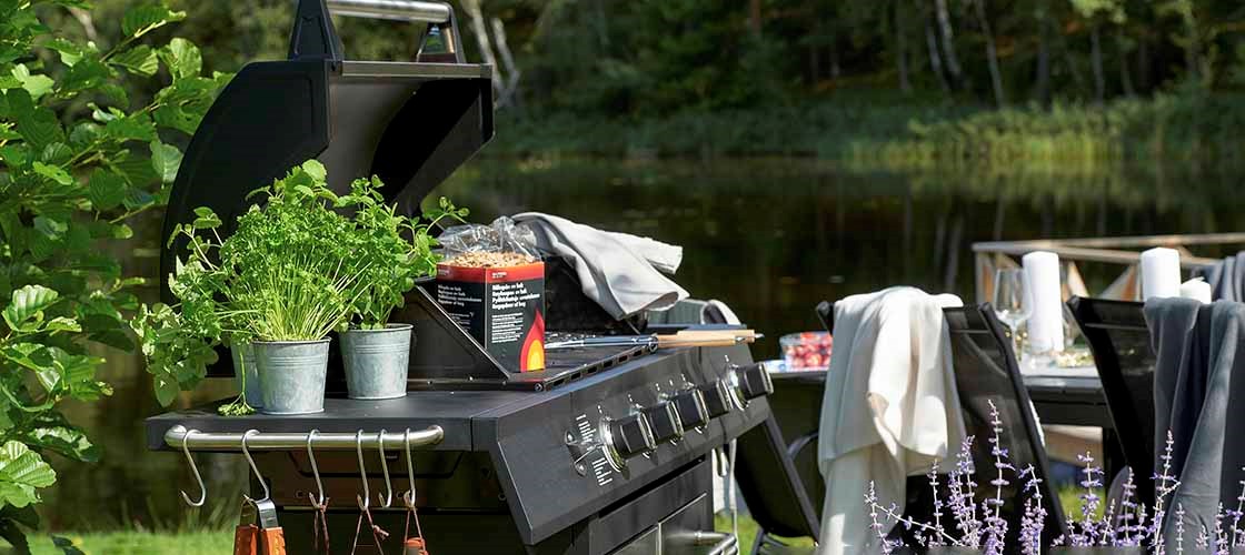 Gasolgrill utomhus vid en sjö, omgiven av grönska. På grillens sidobord står krukor med örter. I bakgrunden syns ett dukat bord och stolar i en trädgårdsmiljö.