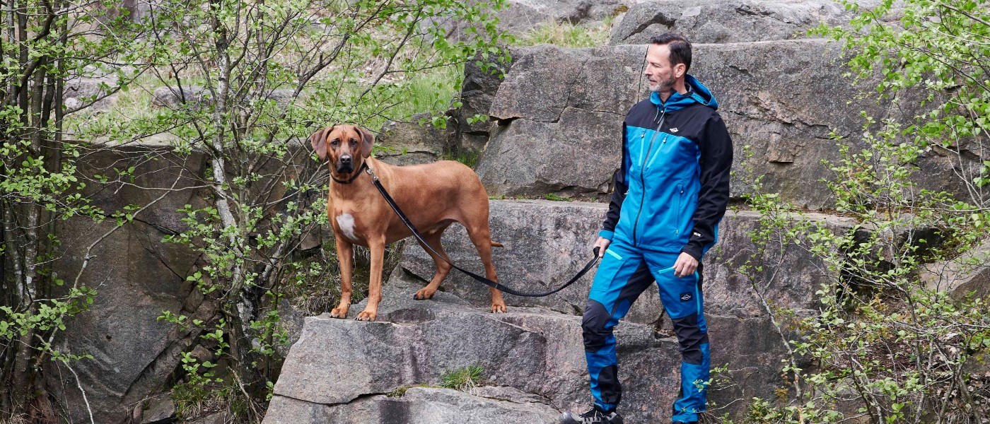 En person i blå friluftsklädsel står på klippor i en grönskande naturmiljö och håller en hund i koppel. Hunden står på en stenavsats och tittar framåt medan kopplet löper mellan hunden och personen. Stora klippblock, buskar och träd omger platsen.