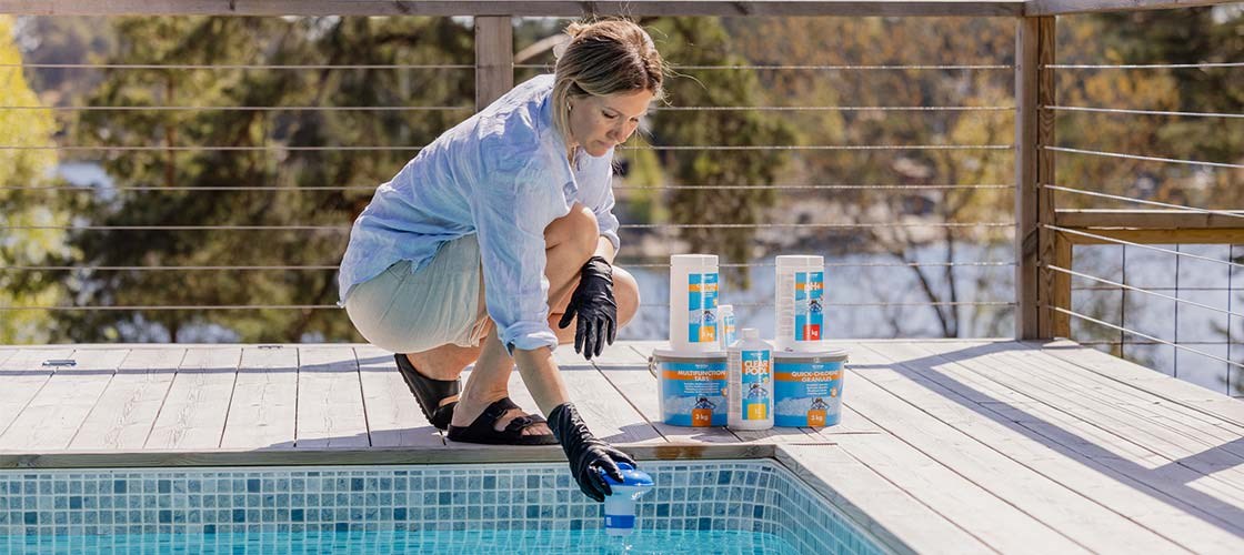 A person crouches at the edge of a swimming pool on a sunlit wooden deck and places a blue dosing device into the water, with several containers of pool chemicals lined up nearby.