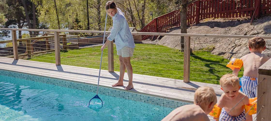 En kvinna står på trädäcket vid en pool och använder en håv i vattnet, medan tre barn sitter vid kanten och leker med färgglada armpuffar på sig. I bakgrunden syns gräsmatta, träd och ett rött staket i solljus.