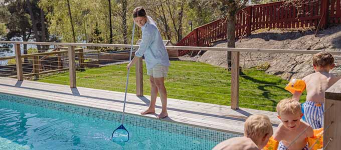 En kvinna står vid poolkanten och rengör poolen med en håv. På sidan av poolen leker tre barn iklädda badkläder och armpuffar.