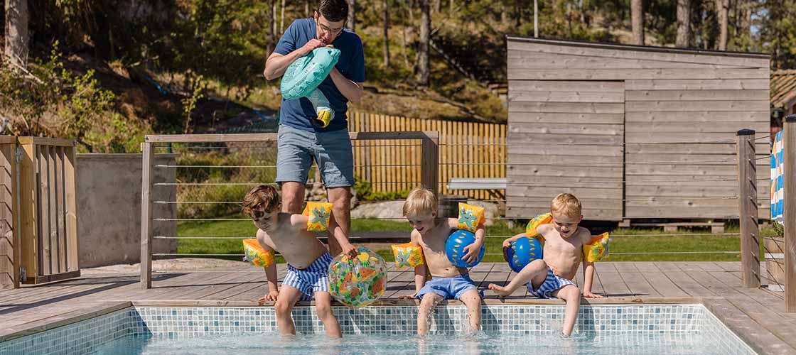 Flera personer sitter och står vid kanten av en utomhuspool. Tre barn doppar fötterna i poolen och en vuxen man står i bakgrunden och blåser upp en badring. I bakgrunden syns ett trädäck, staket, gräsyta och en mindre byggnad omgiven av träd. Bilden visar en sommarmiljö där människor vistas vid en pool för bad och avkoppling.