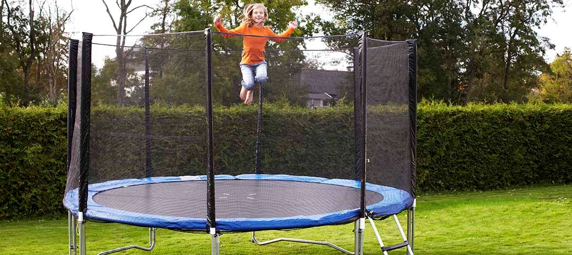 En person hoppar på en rund studsmatta med skyddsnät som står på en gräsmatta i en trädgård. Studsmattan har en stege på sidan och omges av häckar och träd i bakgrunden. Bilden visar en utomhusmiljö med fokus på användning av studsmatta med skyddsnät för ökad säkerhet.