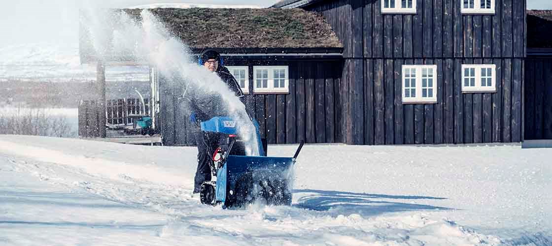 En man klädd i vinterkläder, mössa och hörselskydd kör en blå snöslunga som sprutar snö. I bakgrunden står ett mörkt trähus och det är mycket snö på marken.