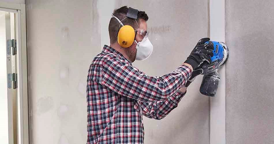 En man som är klädd i rutig arbetsskjorta, arbetshandskar, hörselskydd och munskydd slipar en gipsvägg med en batteridriven, blå slipmaskin.