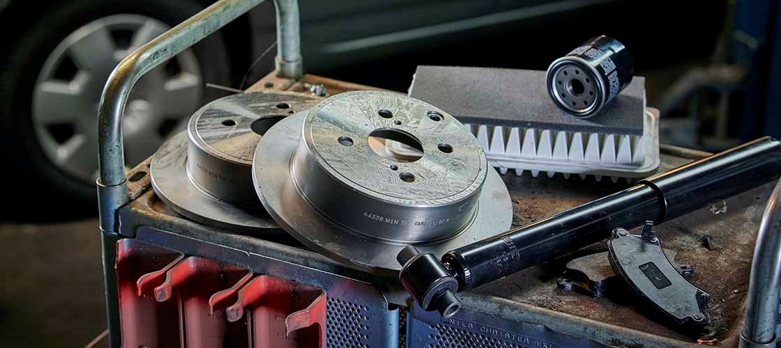 Närbild av en verkstadsvagn med bildelar och verktyg. Två bromsskivor ligger staplade bredvid ett oljefilter, en drivaxel och ett digitalt mätverktyg. I bakgrunden syns delar av ett garage med en bil i oskärpa.