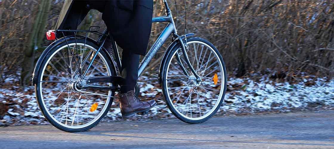 En grå cykel med texten Yosemite på ramen framförs av en person med bruna skor. Det är snö på marken och buskar utan löv i bakgrunden.